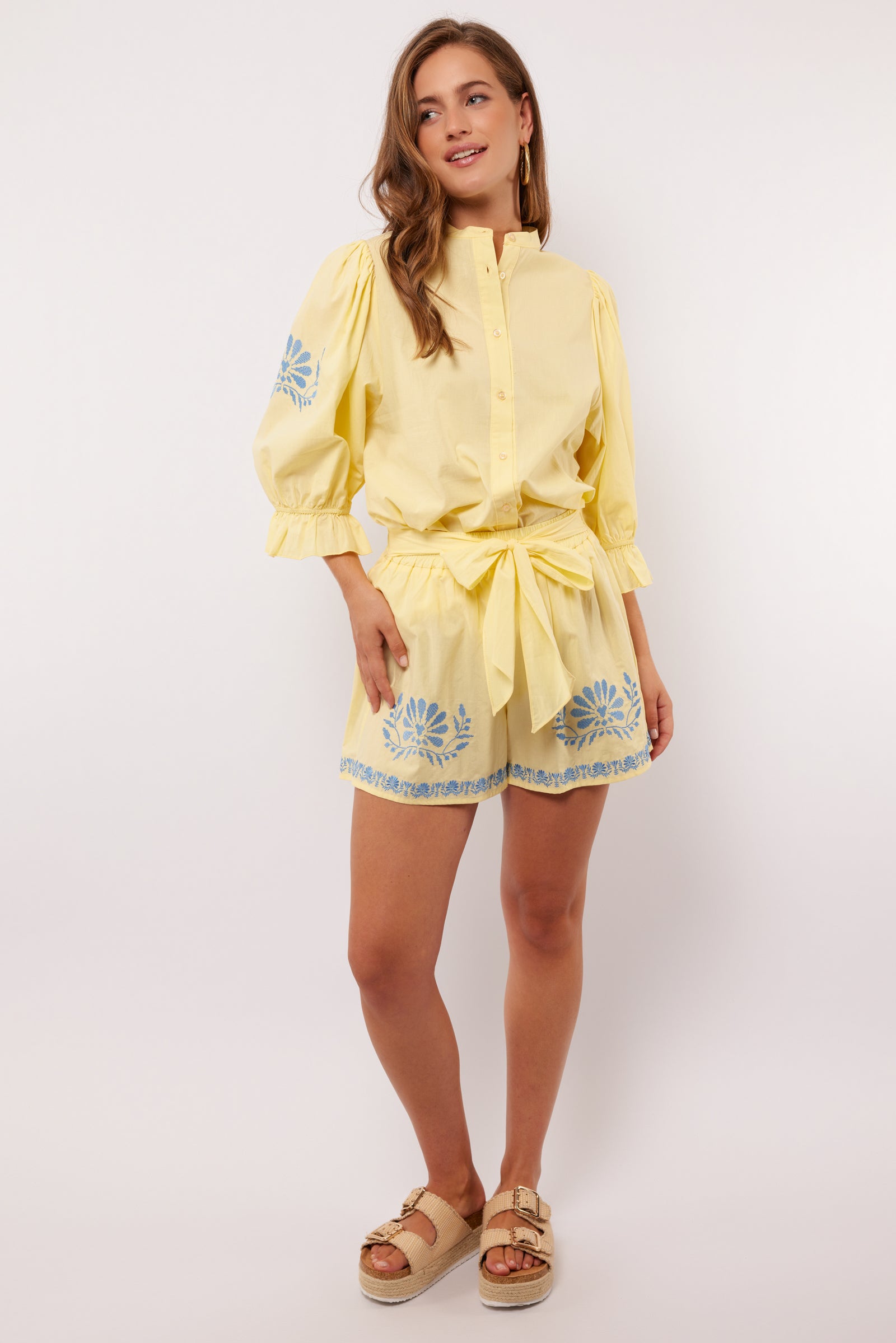 Vrouw draagt gele Fluresk Saine short met blauwe bloemenprint en bijpassende blouse, zomerse outfit met sandalen.