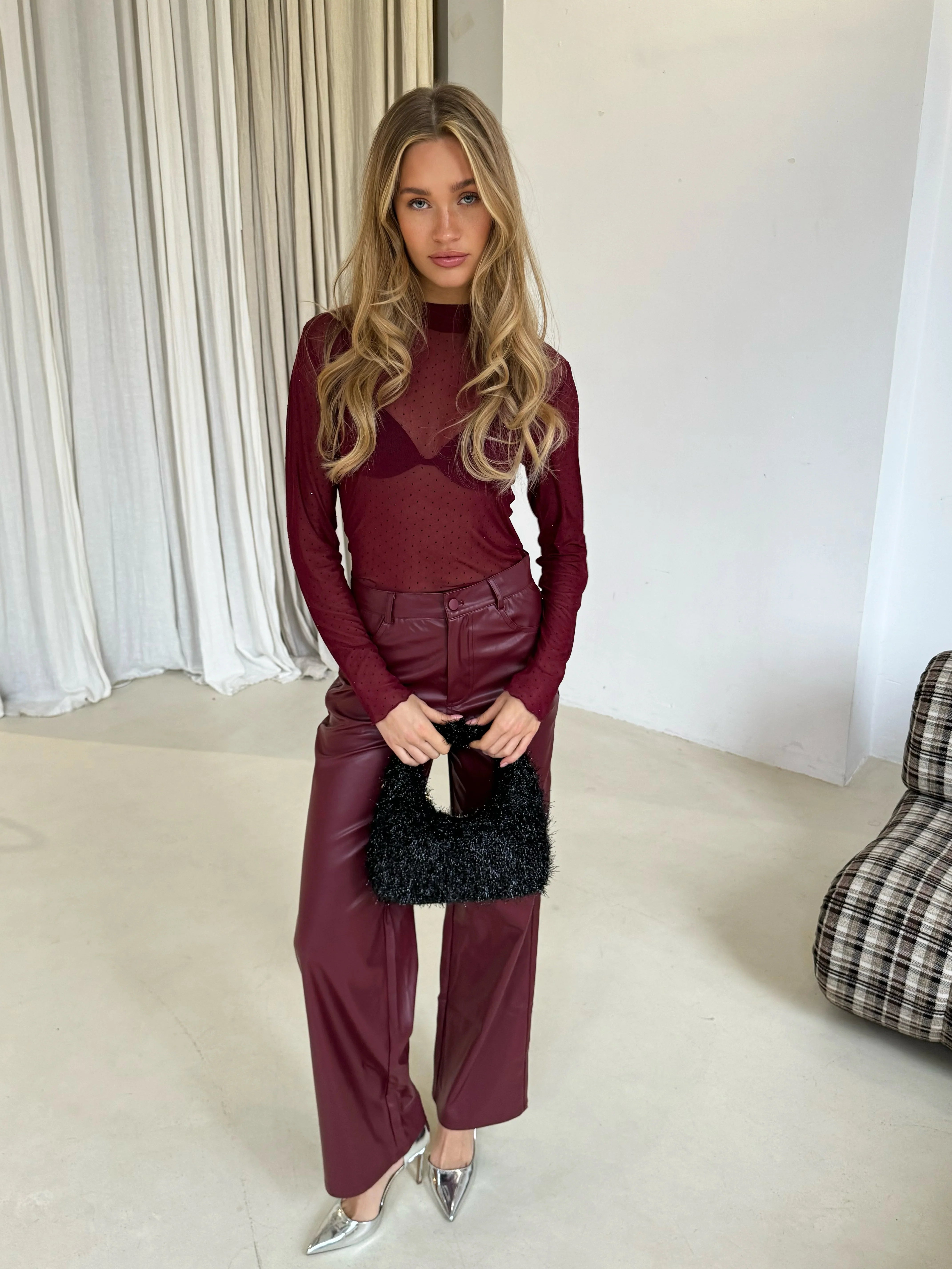 Model draagt een stijlvolle bordeauxrode Ambika-top met doorschijnende stof en strass-steentjes, gecombineerd met bordeauxrode leren broek en zilveren hakken. Ze houdt een zwarte tas met textuur vast