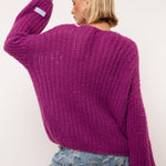 Model draagt de Florin Cardigan van Fluresk in een levendige paarse kleur. De achterkant van de cardigan wordt benadrukt, met een losse pasvorm en zachte gebreide textuur. De cardigan is gecombineerd met lichtblauwe jeans met een ruige afwerking.