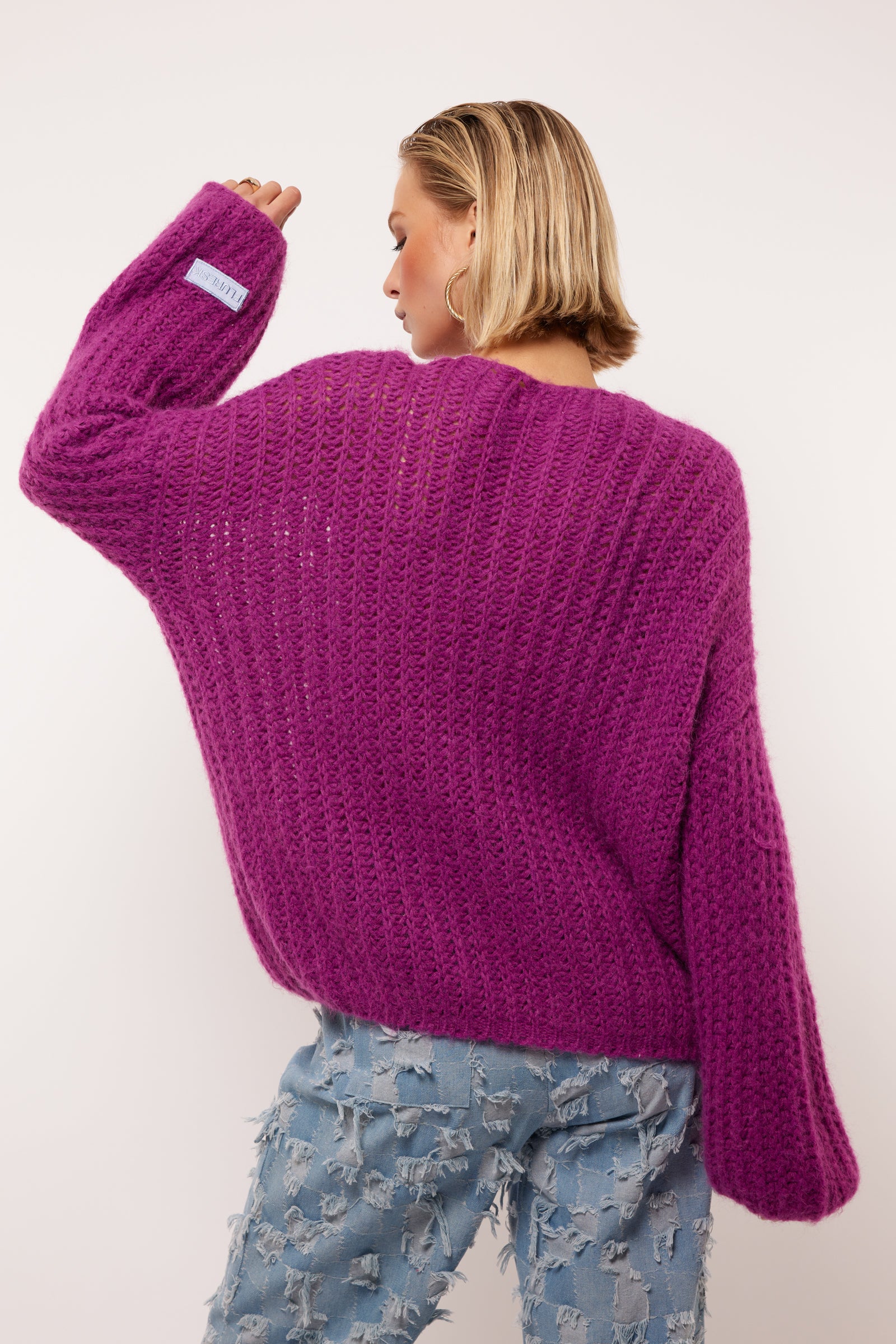 Model draagt de Florin Cardigan van Fluresk in een levendige paarse kleur. De achterkant van de cardigan wordt benadrukt, met een losse pasvorm en zachte gebreide textuur. De cardigan is gecombineerd met lichtblauwe jeans met een ruige afwerking.