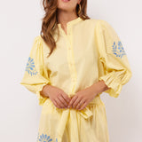Model draagt gele Fluresk Marlowe blouse met bijpassende short en blauwe borduursels.