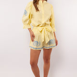 Vrouw draagt gele Fluresk Saine short met blauwe bloemenprint en bijpassende blouse, zomerse outfit met sandalen.