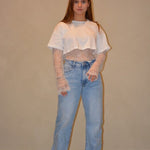 Model draagt een witte cropped top gecombineerd met een kanten top eronder en een lichtblauwe jeans met een casual pasvorm, gestyled met zwarte schoenen.