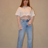 Model draagt een witte cropped top gecombineerd met een kanten top eronder en een lichtblauwe jeans met een casual pasvorm, gestyled met zwarte schoenen.