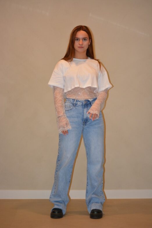 Model draagt een witte cropped top gecombineerd met een kanten top eronder en een lichtblauwe jeans met een casual pasvorm, gestyled met zwarte schoenen.